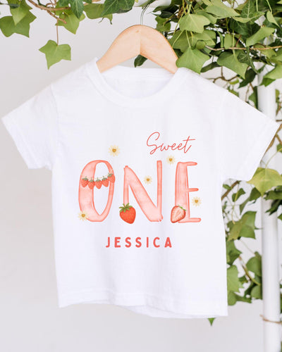 A white t-shirt with a personalized text and design printed on it, hanging on a wooden hanger.