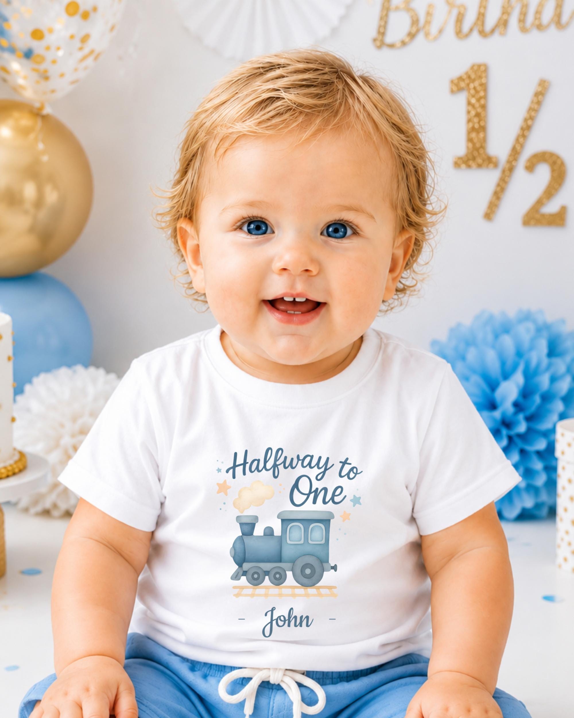 A smiling baby boy wearing a white t-shirt with a train graphic and the text "Halfway to One" is sitting in front of a birthday party backdrop with gold and blue decorations.