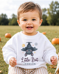 A smiling baby boy wearing a white sweatshirt with a bat graphic and the text "Little Bat" and "Johnson" printed on it, sitting in a field with pumpkins in the background.