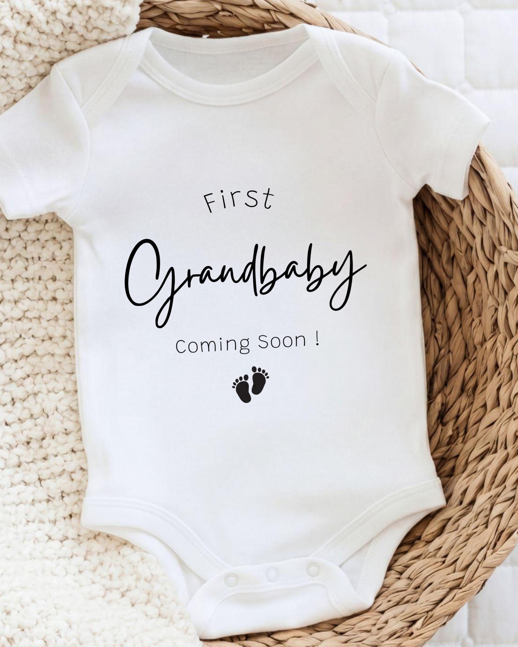 A white onesie with the text "First Grandbaby Coming Soon!" printed on it, along with a small footprint icon.