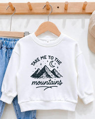 A white sweatshirt with a graphic design of mountains and stars, along with the text "Take me to the mountains" printed on it, hanging on a wooden hanger.