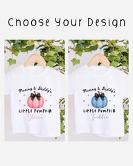 two white t-shirts with personalized text and illustrations, hanging on a wooden hanger. The text on the t-shirts reads "Mummy & Daddy's LITTLE PUMPKIN Olivia" and "Mummy & Daddy's LITTLE PUMPKIN Freddie", indicating that the t-shirts are designed for a mother and father to wear. The t-shirts are set against a backdrop of greenery, creating a natural and serene atmosphere.