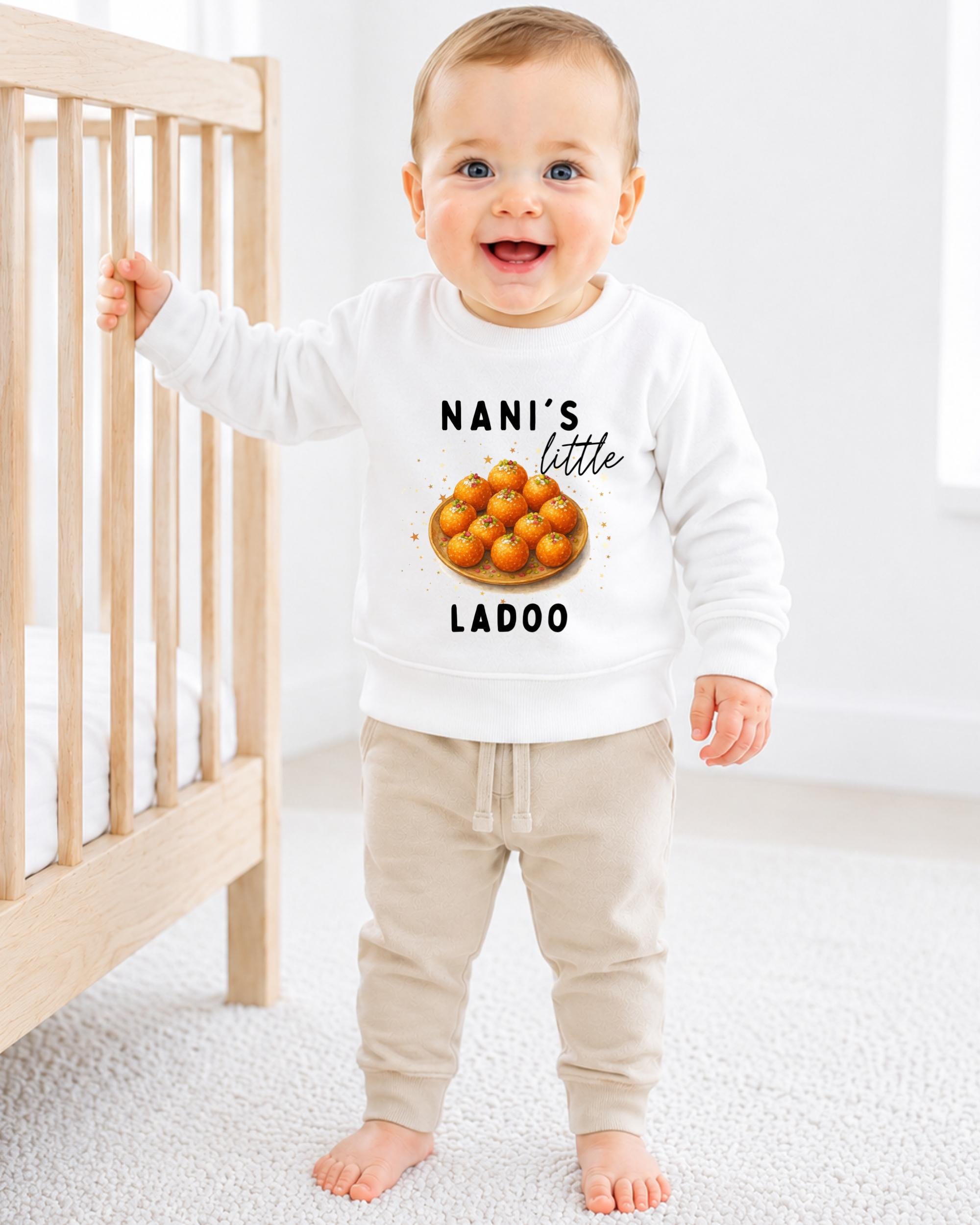 A smiling baby boy wearing a white sweatshirt with a graphic of oranges and the text "NANI'S LITTLE LADOO" standing in a nursery.