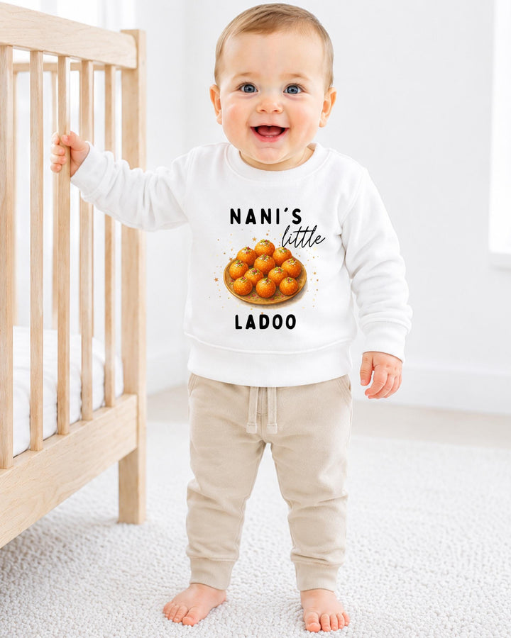 A smiling baby boy wearing a white sweatshirt with a graphic of oranges and the text "NANI'S LITTLE LADOO" standing in a nursery.