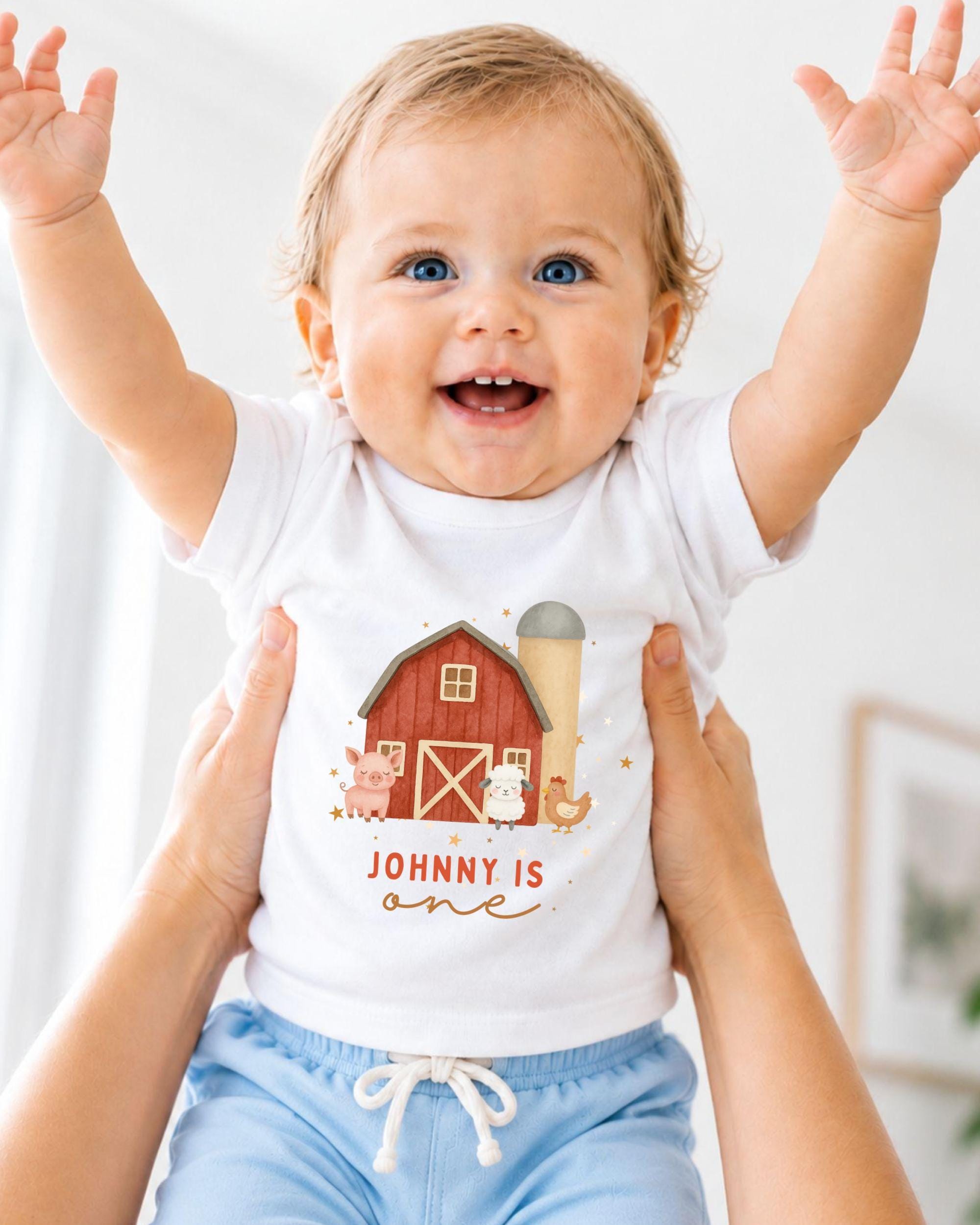 A smiling baby wearing a white t-shirt with a farm scene and the name "Johny" printed on it, being held up by someone's arms.