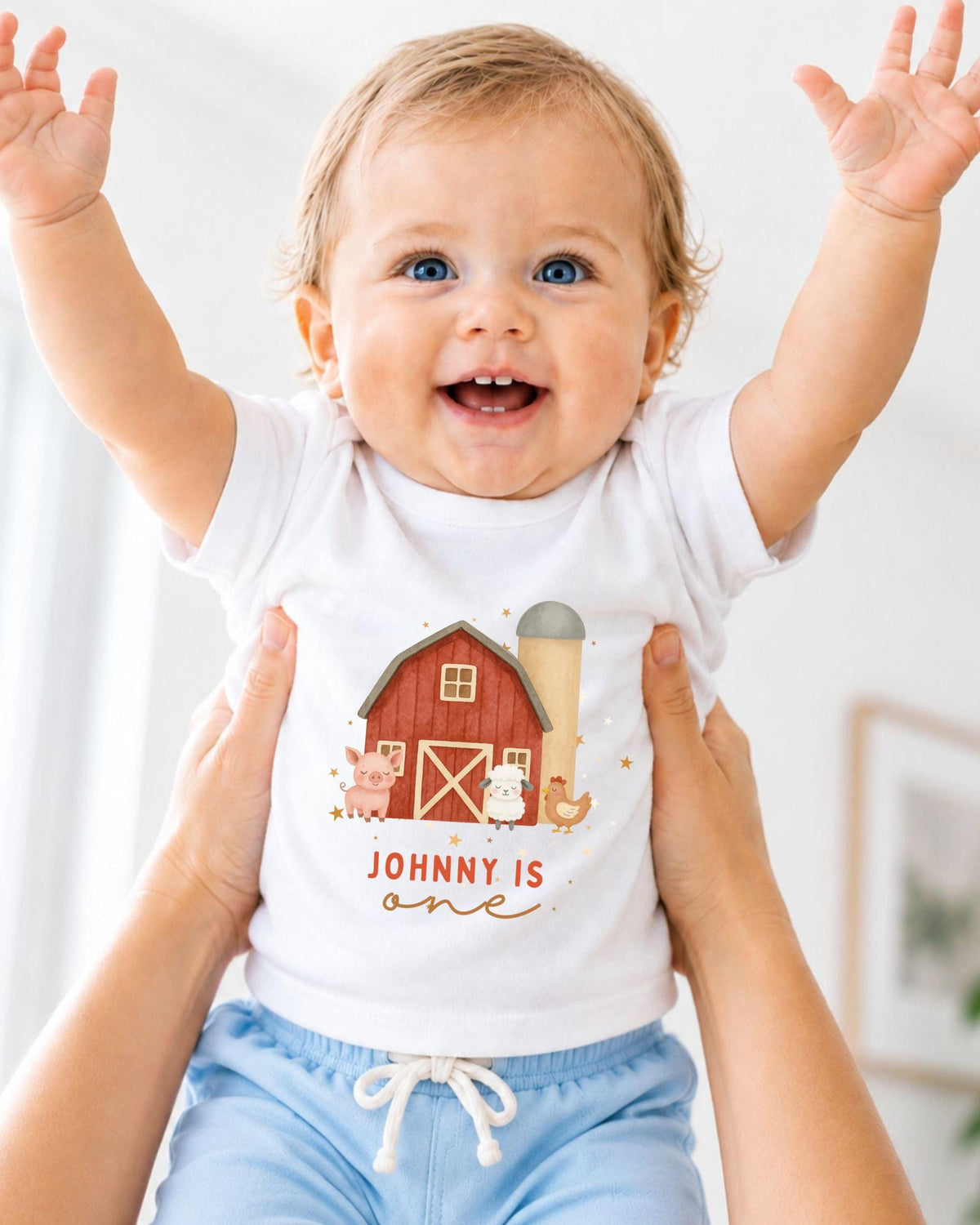 A smiling baby wearing a white t-shirt with a farm scene and the name "Johny" printed on it, being held up by someone's arms.
