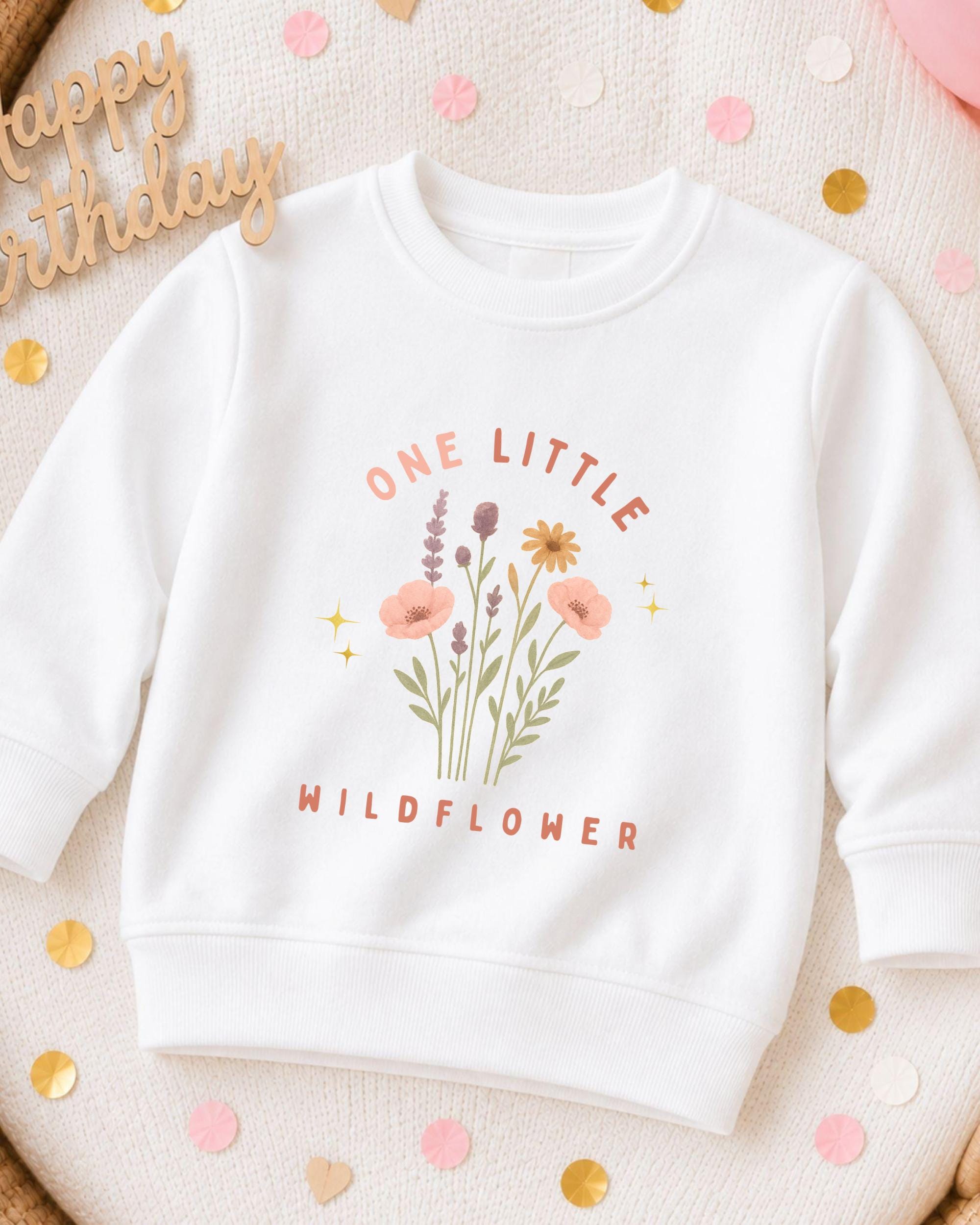A white sweatshirt with a floral design and the text "One Little Wildflower" printed on it, surrounded by confetti.