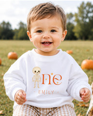 A smiling baby boy wearing a white sweater with a skeleton and star design, sitting in a field with pumpkins in the background.
