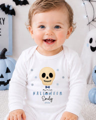 A smiling baby boy wearing a white onesie with a skull and snowflake design, sitting in front of Halloween decorations.