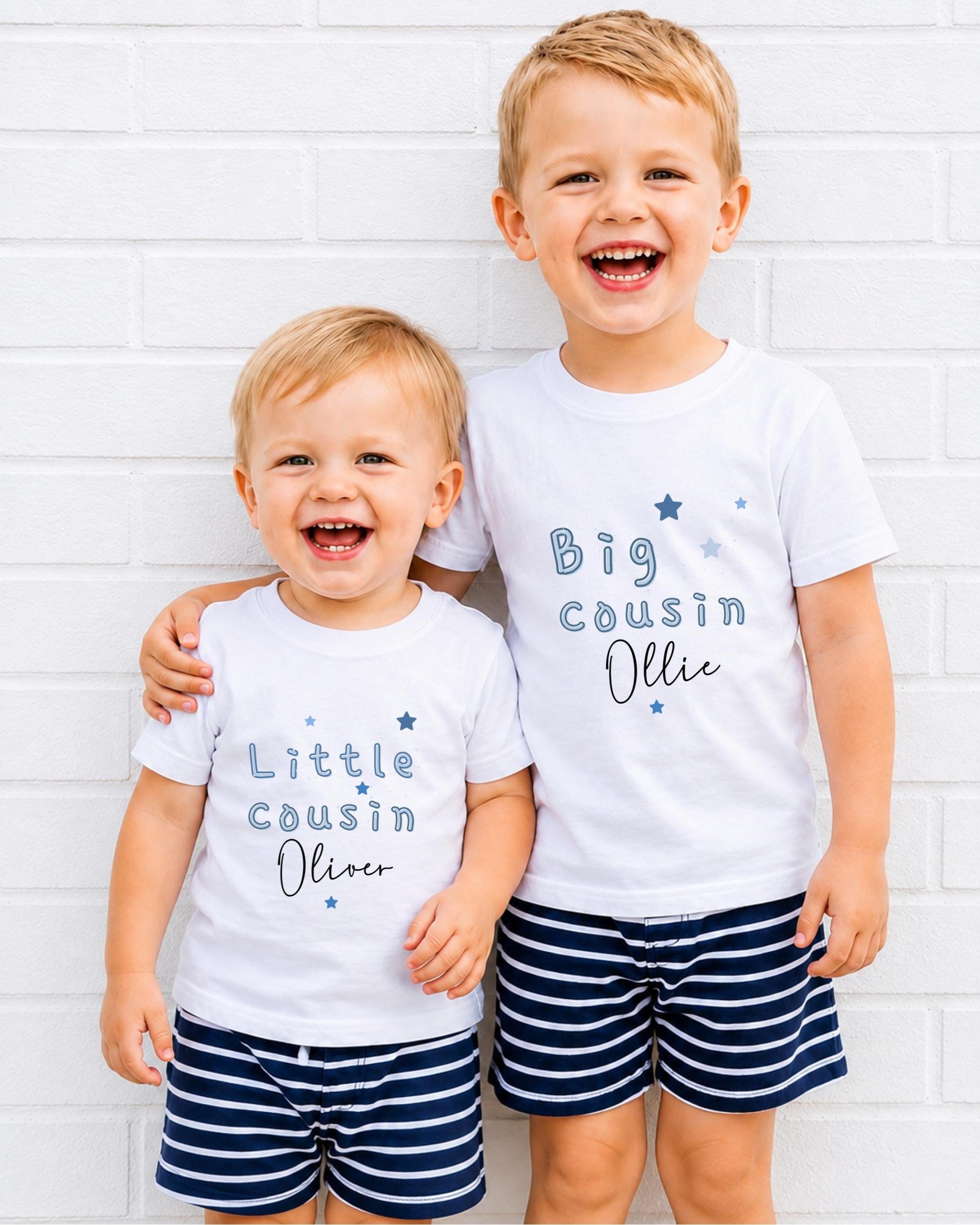 Two young boys wearing matching white t-shirts with text and stars, smiling and posing together against a white brick wall.