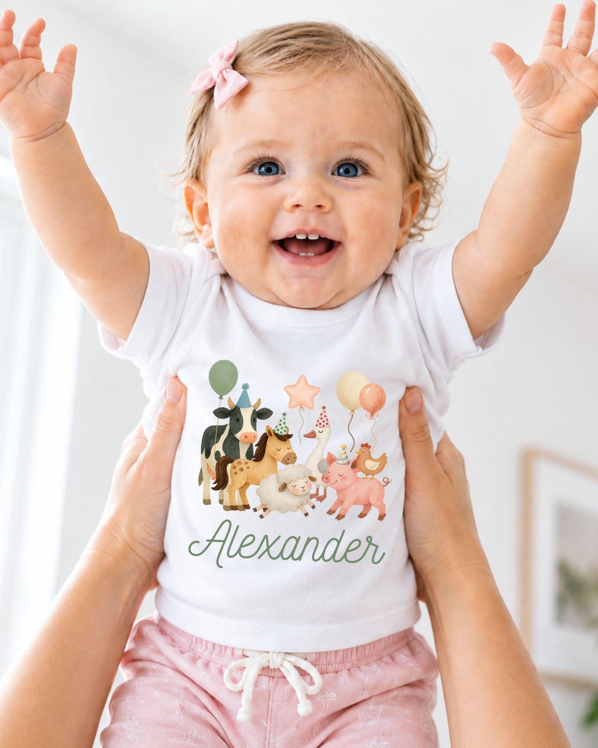A smiling baby girl wearing a white t-shirt with a cartoon animal design and the name "Alexander" printed on it, being held up by someone's arms.