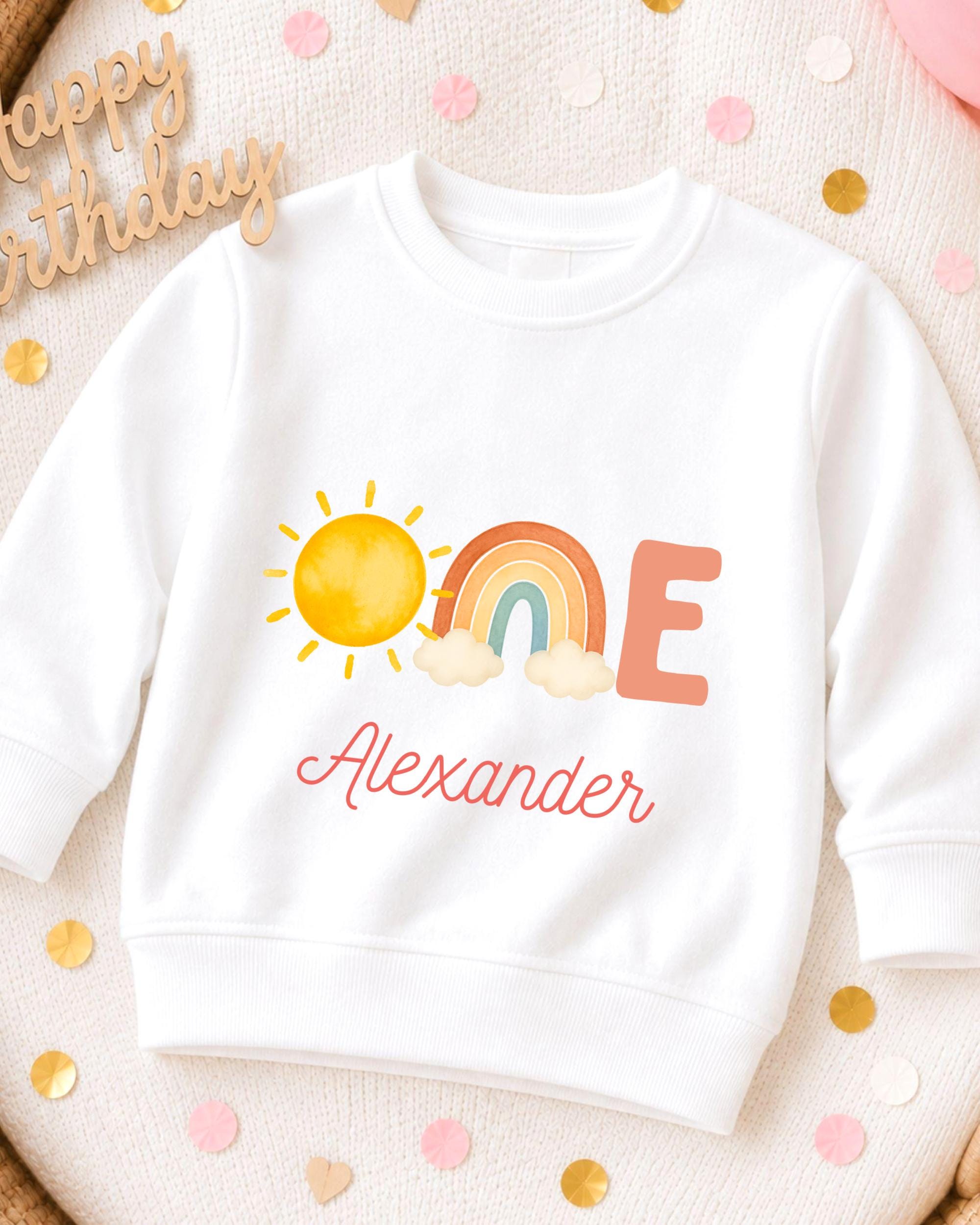 A white sweatshirt with a colorful graphic design featuring a sun, rainbow, and clouds, along with the name "Alexander" written on it.