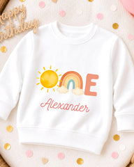 A white sweatshirt with a colorful graphic design featuring a sun, rainbow, and clouds, along with the name "Alexander" written on it.