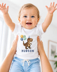 A smiling baby wearing a white t-shirt with a dinosaur graphic and the name "HUDSON" printed on it, being held up by someone's arms.
