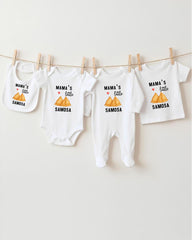 A row of four white baby clothes with the text "MAMA'S LITTLE SAMOSA" printed on them, hanging on a clothesline.