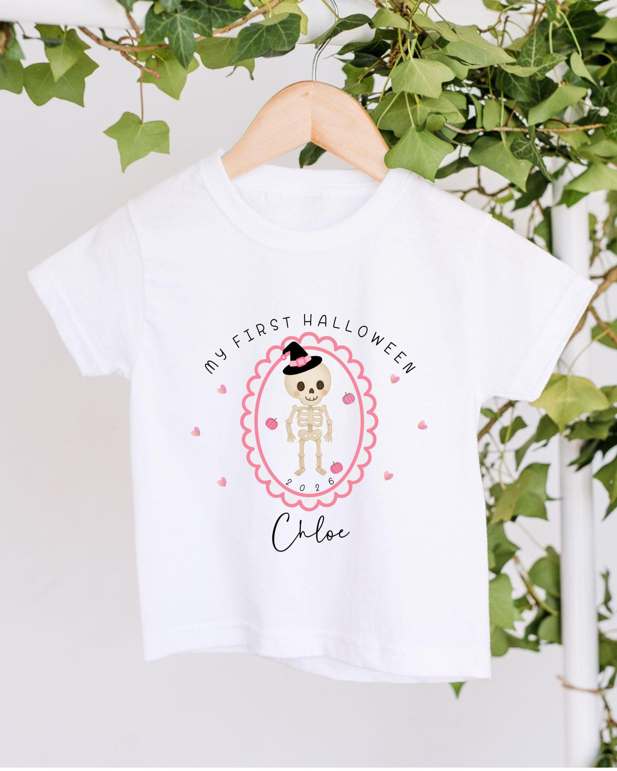 A white t-shirt with a Halloween-themed design hanging on a wooden hanger. The design features a skeleton wearing a witch's hat and surrounded by pink hearts.