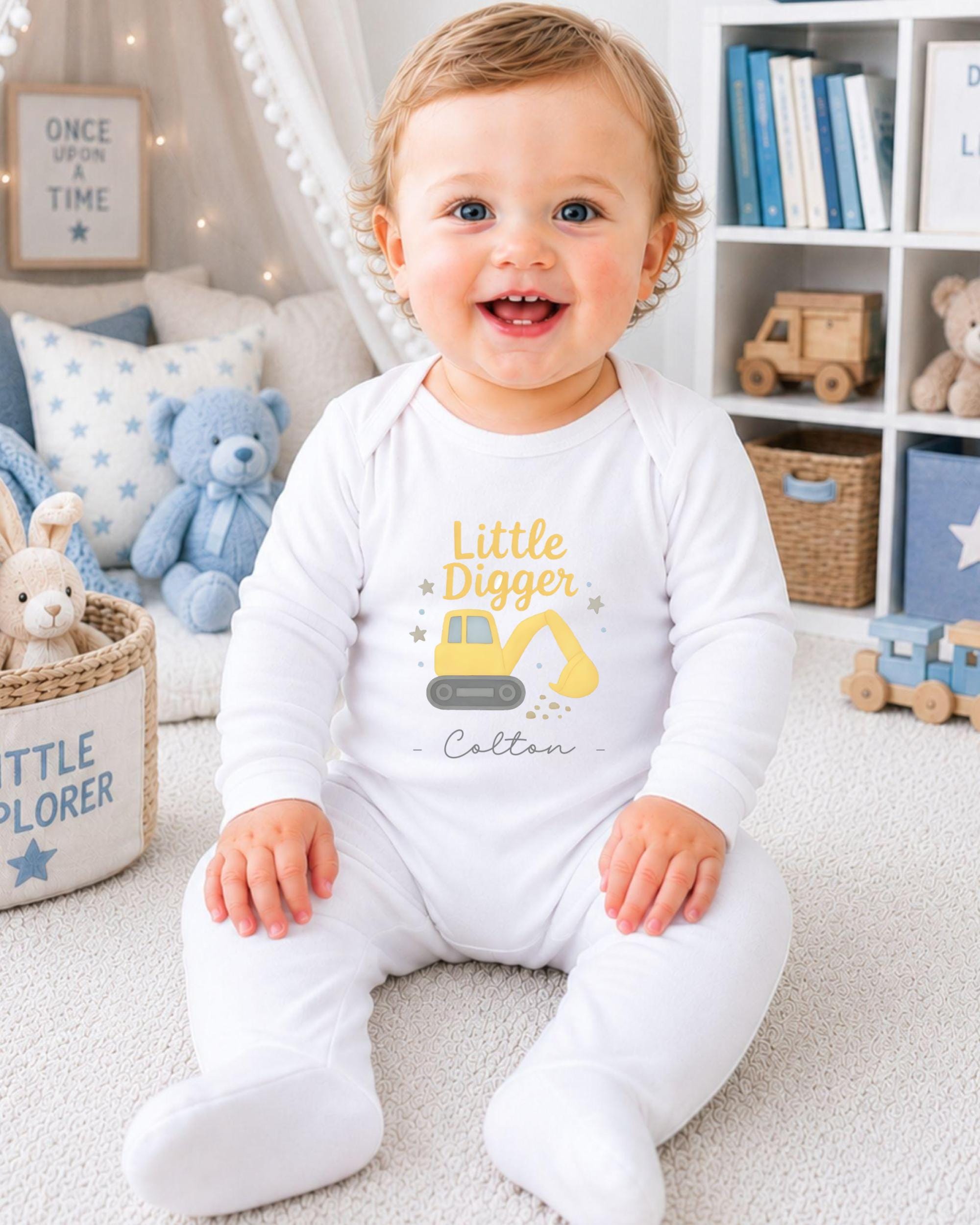 A smiling baby boy wearing a white onesie with a yellow construction vehicle design, sitting on a carpeted floor in a nursery setting with various toys and books around him.