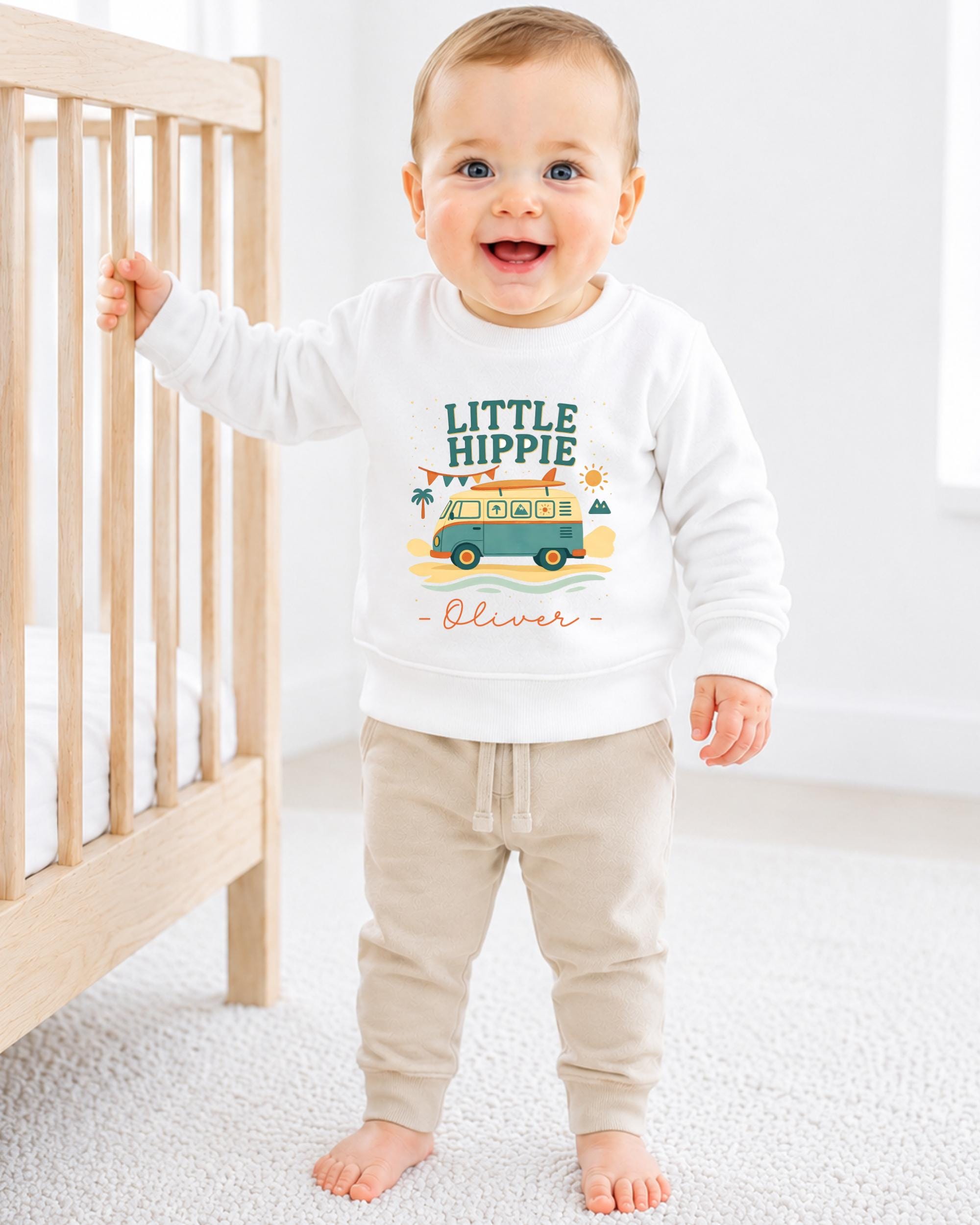 A smiling baby boy wearing a white sweatshirt with a graphic design and the name "Olivier" printed on it, standing in front of a wooden crib.