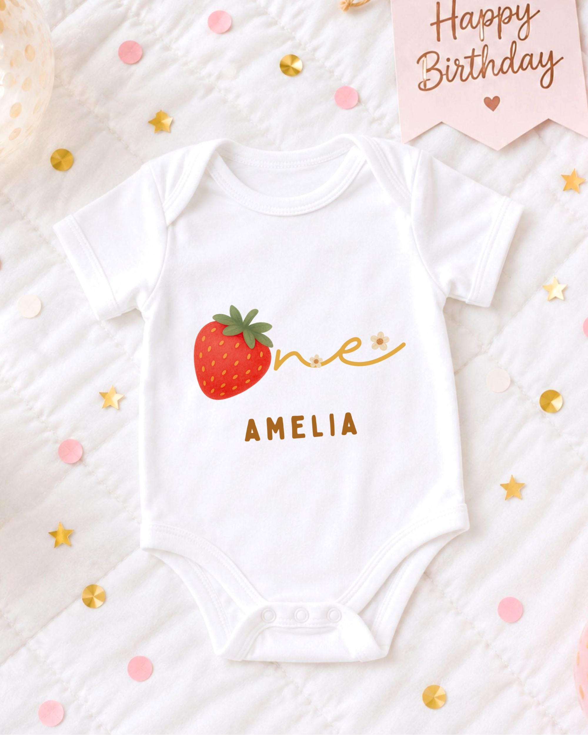 A white onesie with a red strawberry and the name "Amelia" printed on it, surrounded by confetti and a birthday card.