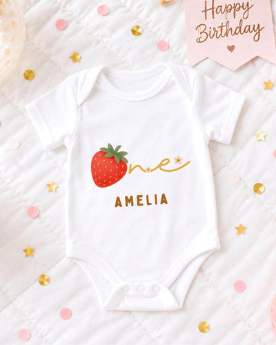 A white bodysuit with a red strawberry and the name "Amelia" printed on it, surrounded by confetti and a birthday card.