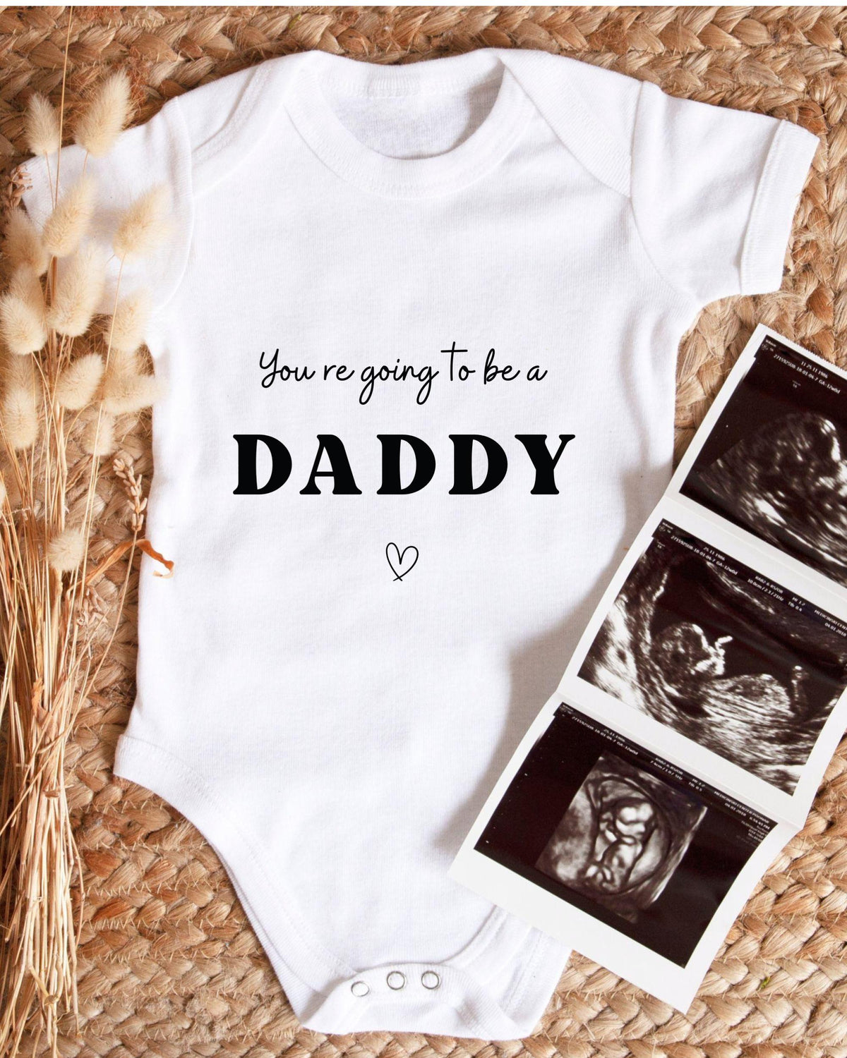 A white baby onesie with the text "You're going to be a DADDY" printed on it, along with a heart symbol. The onesie is placed next to a set of three black and white images depicting an ultrasound image of a baby.