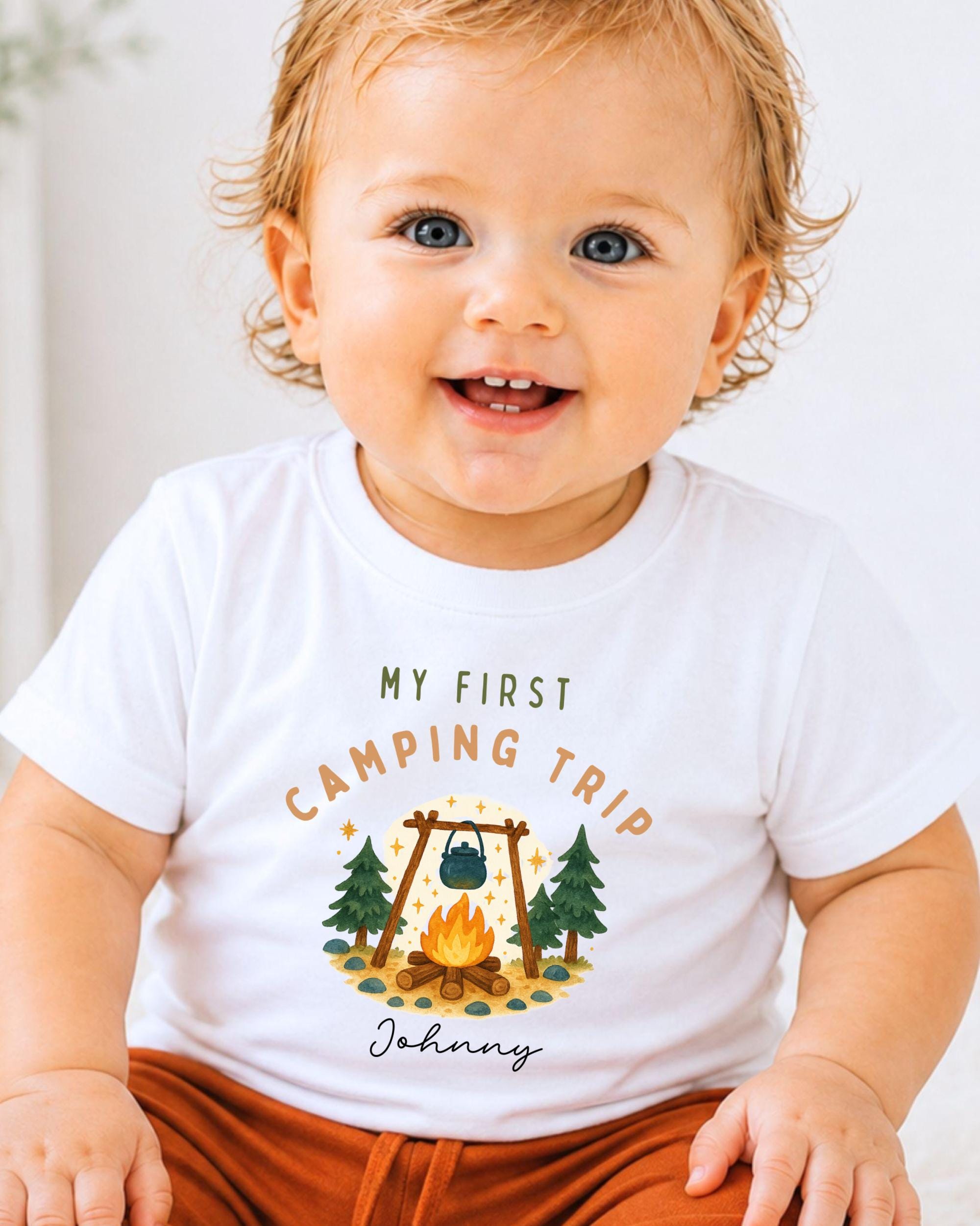A smiling baby wearing a white t-shirt with a camping-themed graphic and text.