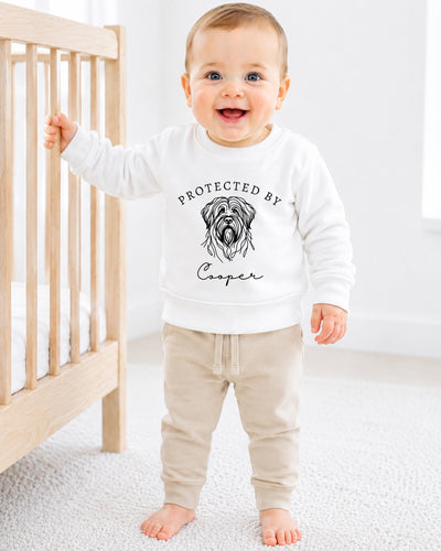 A smiling baby boy wearing a white sweatshirt with a graphic of a dog and the text "PROTECTED BY Cooper" stands next to a wooden crib.