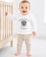 A smiling baby boy wearing a white sweatshirt with a graphic of a dog and the text "PROTECTED BY Cooper" stands next to a wooden crib.