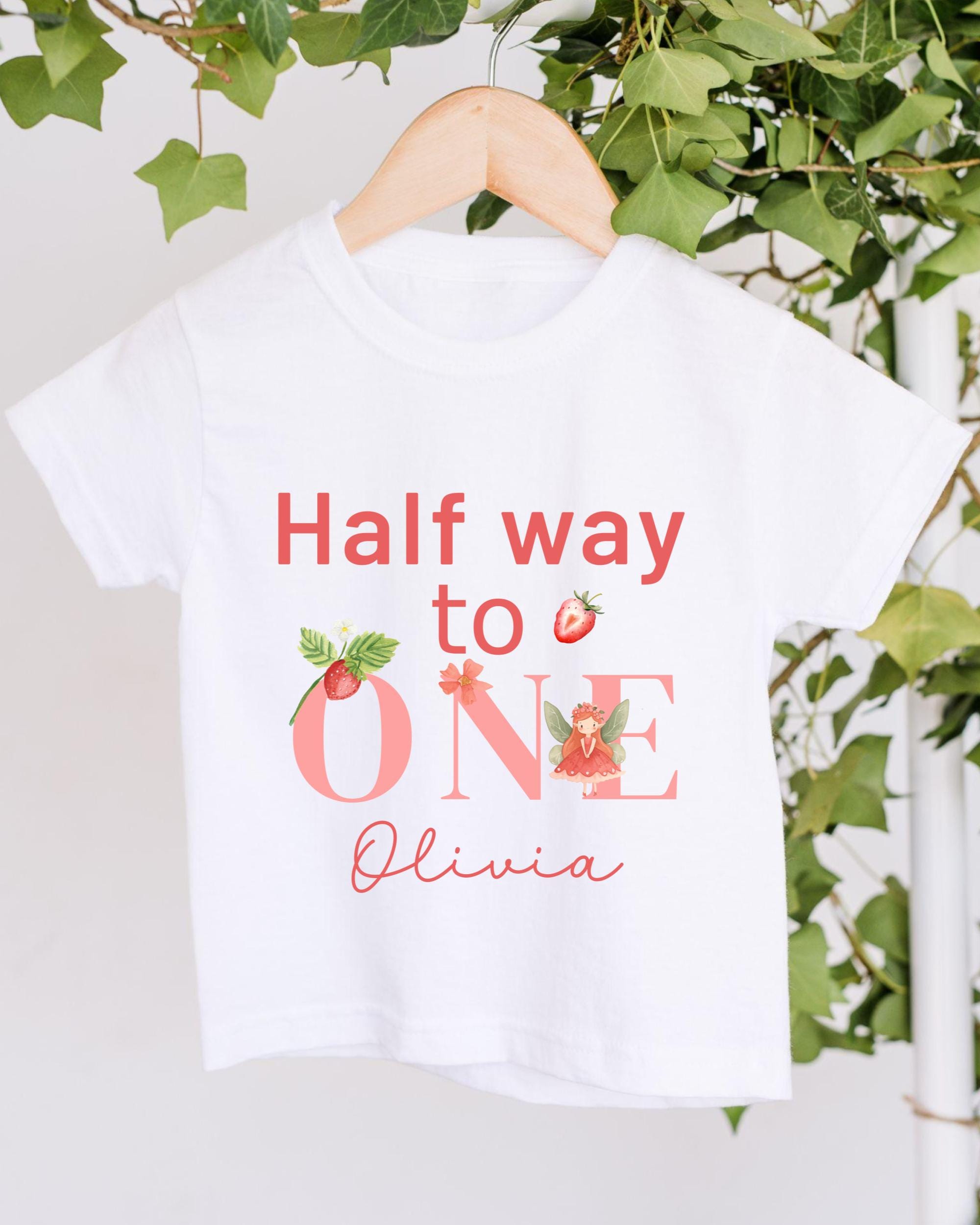 A white t-shirt with the text "Half way to one" and "Olivia" printed on it, along with illustrations of strawberries and a fairy.