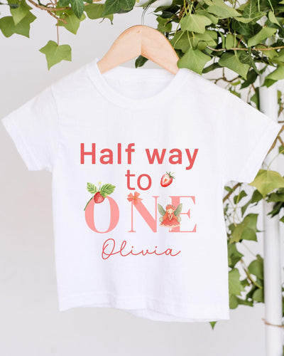 A white t-shirt with the text "Half way to one" and "Olivia" printed on it, along with illustrations of strawberries and a fairy.