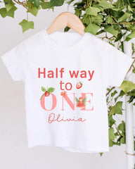 A white t-shirt with the text "Half way to one" and "Olivia" printed on it, along with illustrations of strawberries and a fairy.
