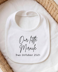 A white bib with the text "Our little Miracle Due October 2025" printed on it.