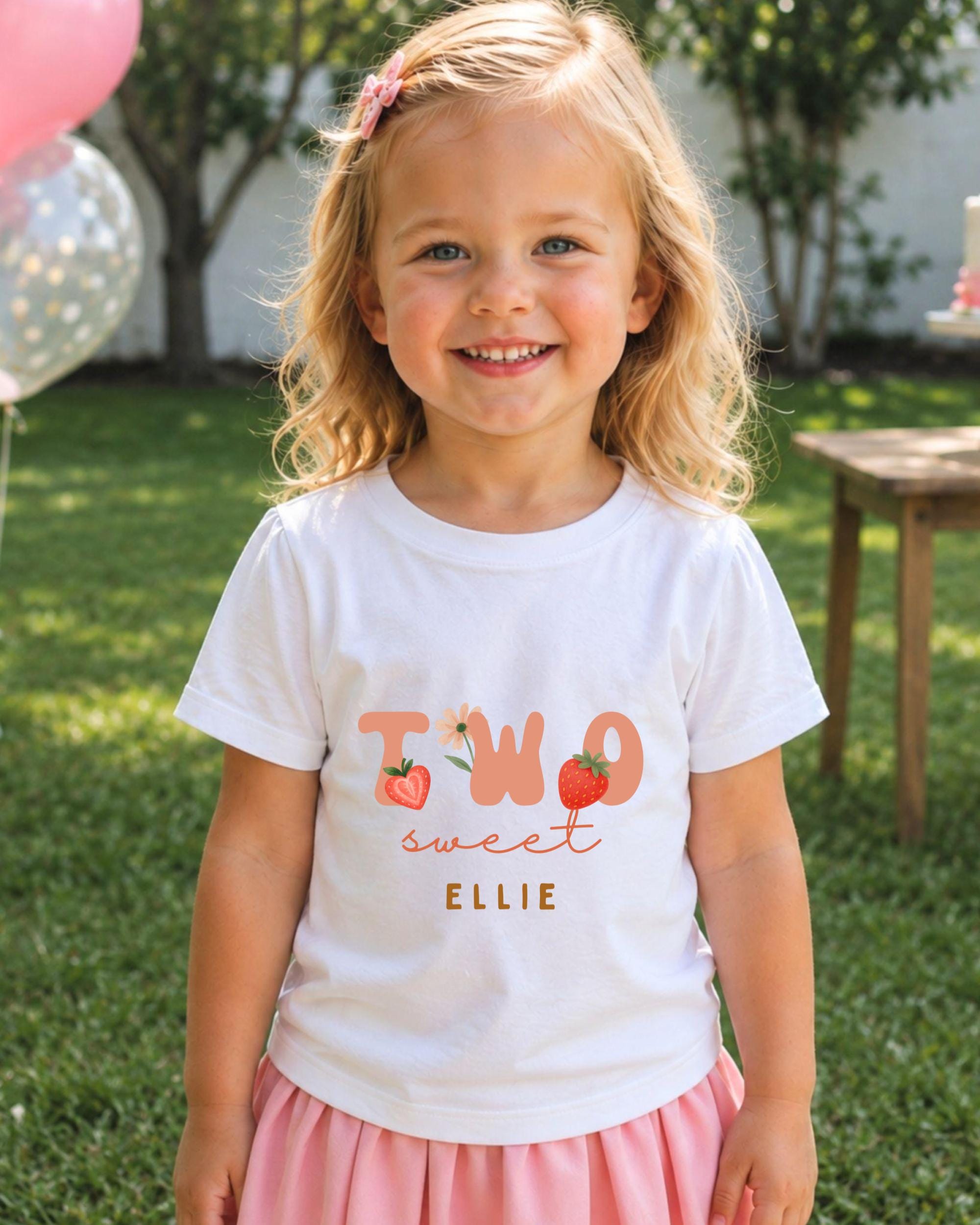 A young girl with blonde hair is smiling and wearing a white t-shirt with the text "Two sweet Ellie" printed on it.