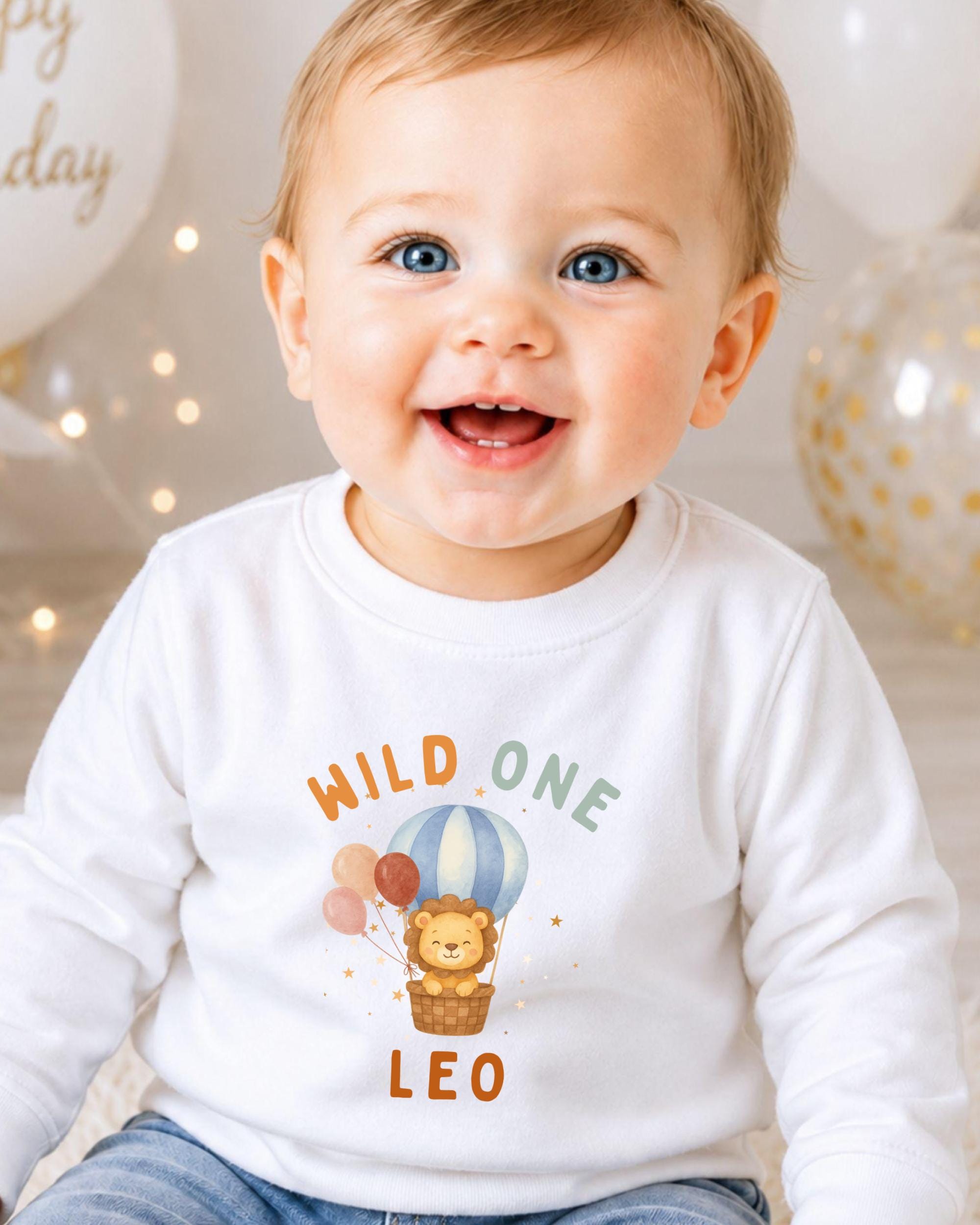 A smiling baby boy wearing a white shirt with a cartoon lion and a hot air balloon graphic, celebrating his first birthday.