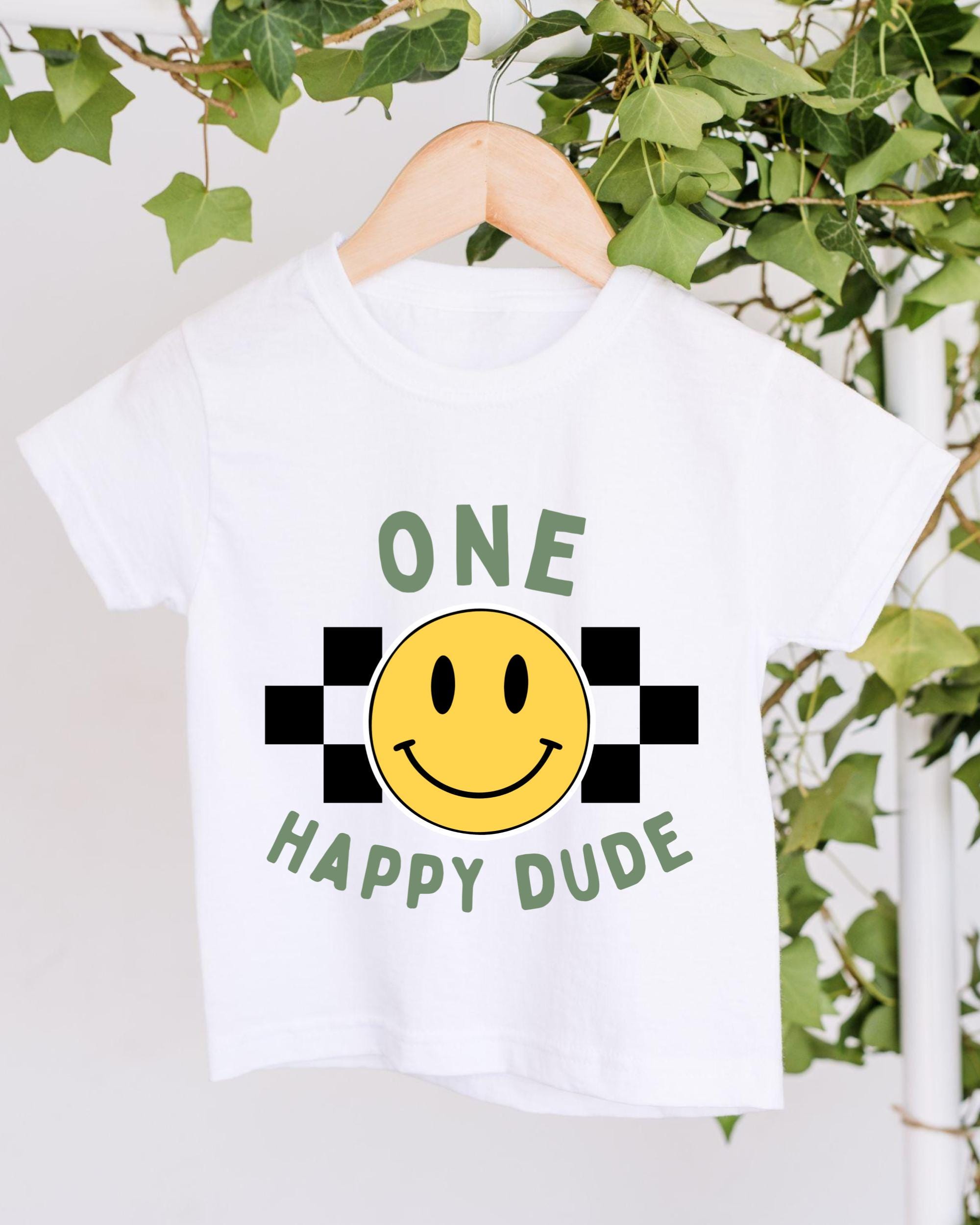 A white t-shirt with a yellow smiley face and the text "ONE HAPPY Dude" printed on it, hanging on a wooden hanger.