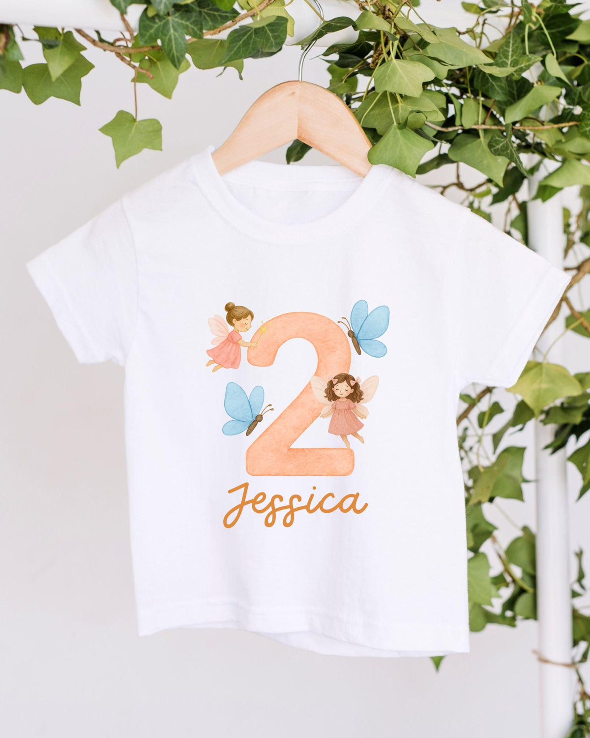 A white t-shirt with a personalized design hanging on a wooden hanger, featuring a large number "2" with two small fairy characters and the name " Jessica" written below.