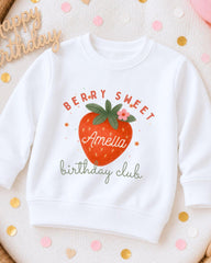 A white sweatshirt with a strawberry graphic and text, along with confetti and a "Happy Birthday" sign.
