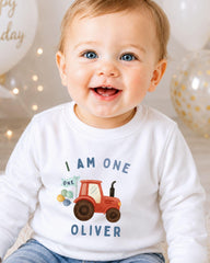A smiling baby boy wearing a white shirt with a tractor graphic and the text "I am one Oliver" printed on it.