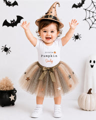 A young girl wearing a witch's hat and a tutu with stars, smiling and raising her arms, standing in front of Halloween decorations.