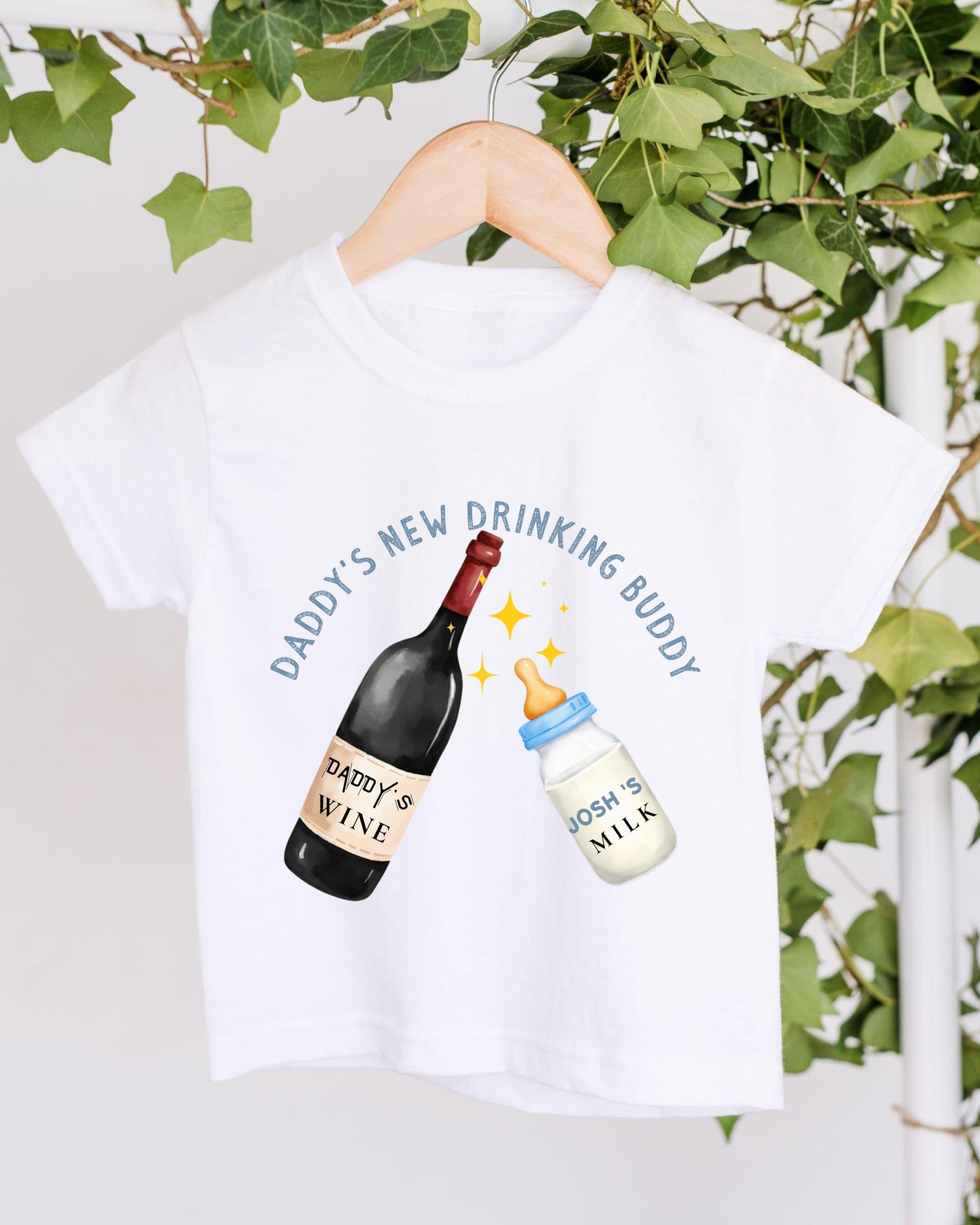 A white t-shirt with a graphic design featuring a bottle of wine and a baby bottle, along with the text "DADY'S NEW DRINKING BUDDY" and "JOSH'S MILK" printed on it.