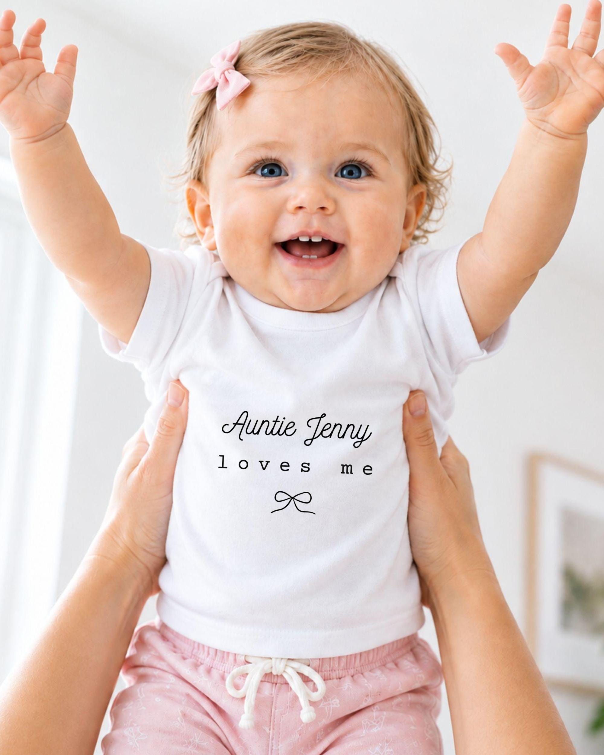 A smiling baby girl wearing a white t-shirt with the text "Auntie Jenny loves me" is being held up by someone's arms.