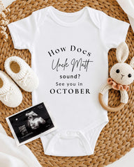 A white baby onesie with text, a crocheted bunny toy, and a baby bootie are arranged on a woven mat.