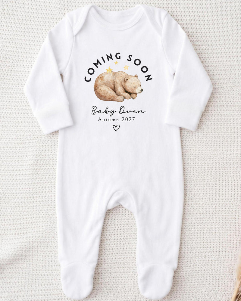 Personalisable white Baby Grow with a sleeping brown watercolour bear beneath curved black 'Coming Soon' lettering, 'Baby Owen Autumn 2027' text and an outlined heart.