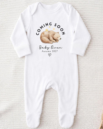 Personalisable white Baby Grow with a sleeping brown watercolour bear beneath curved black 'Coming Soon' lettering, 'Baby Owen Autumn 2027' text and an outlined heart.