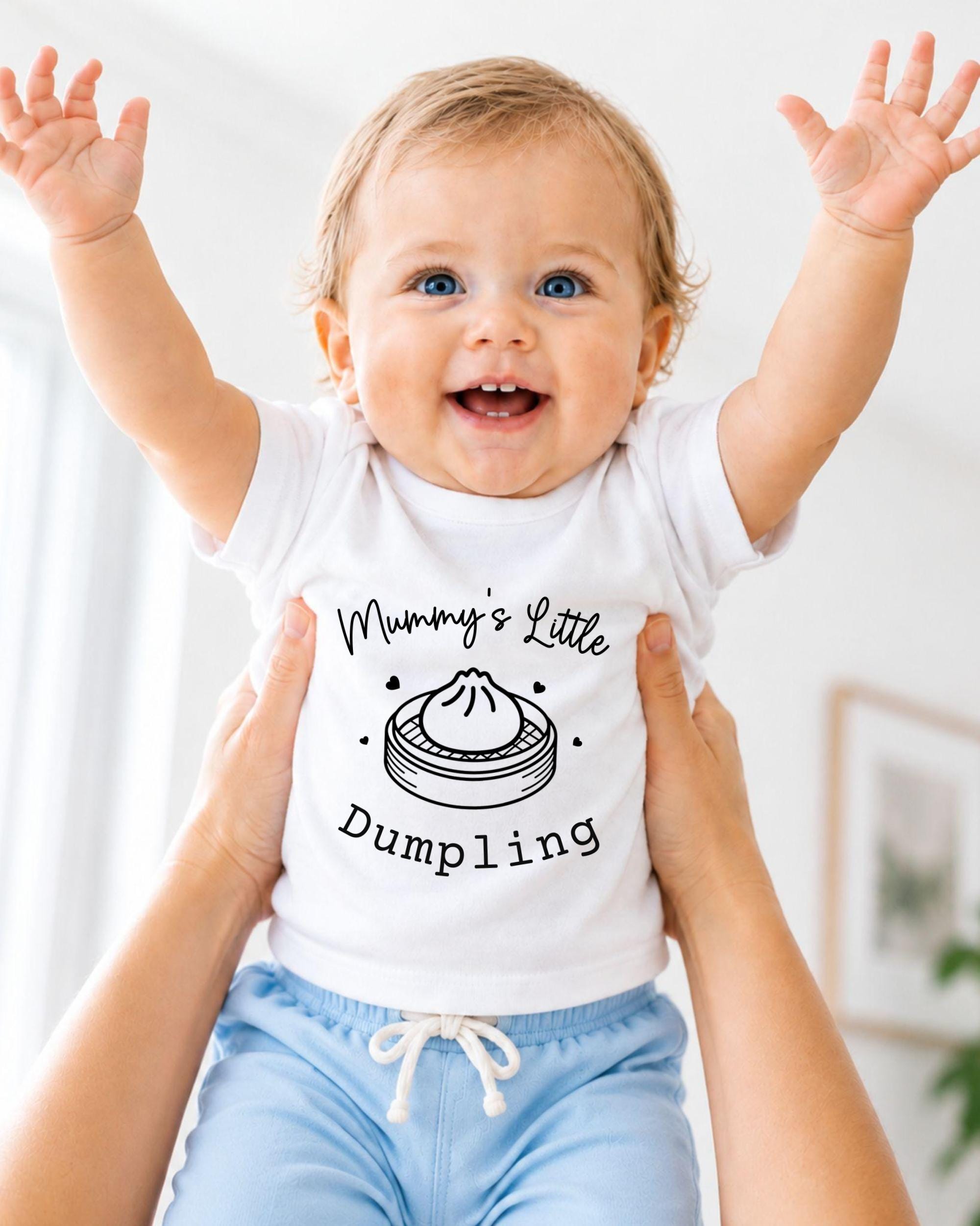 A smiling baby wearing a white t-shirt with a graphic and text, being held up by someone's arms.