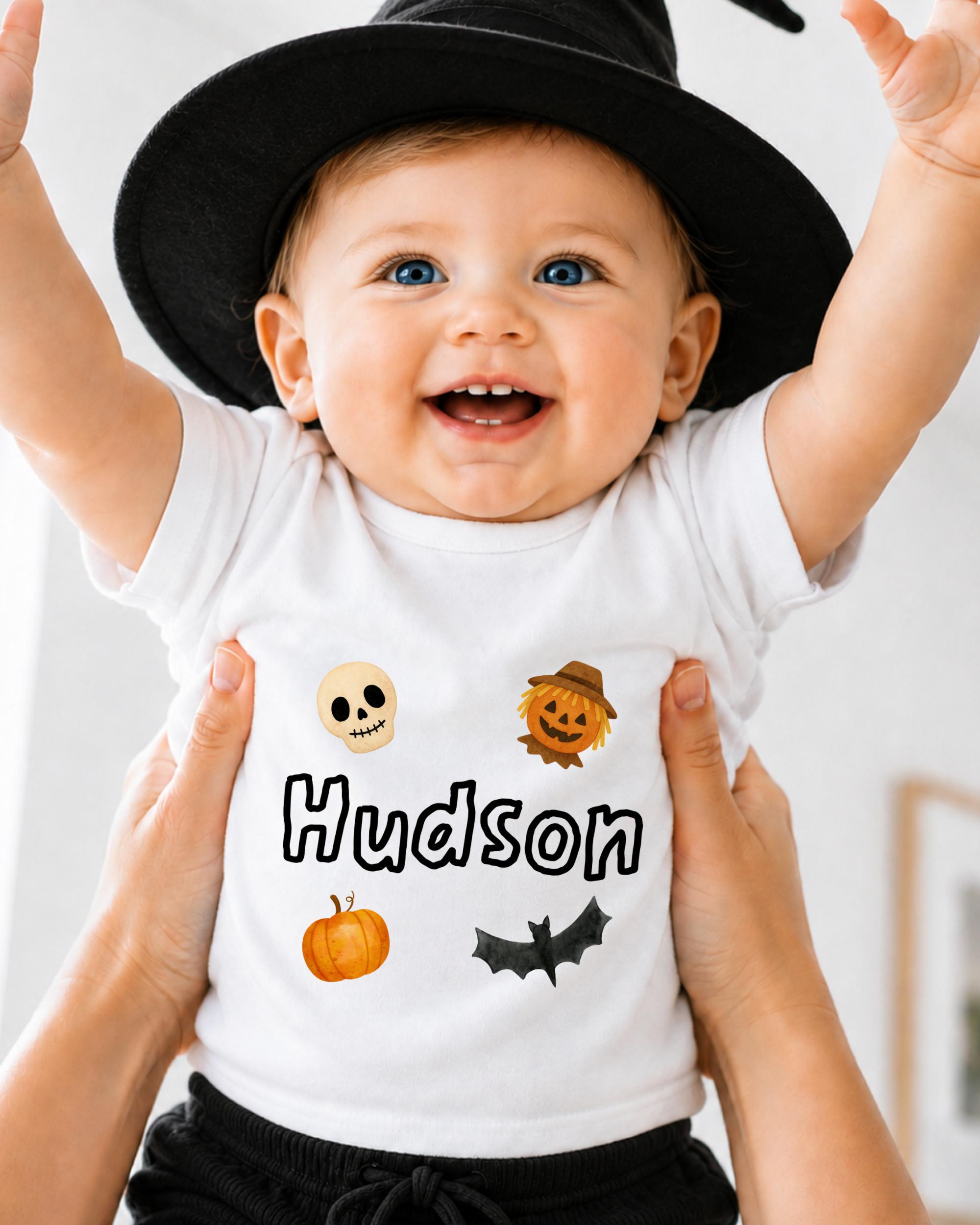 A smiling baby wearing a white t-shirt with Halloween-themed designs and the name "Hudsons" printed on it.