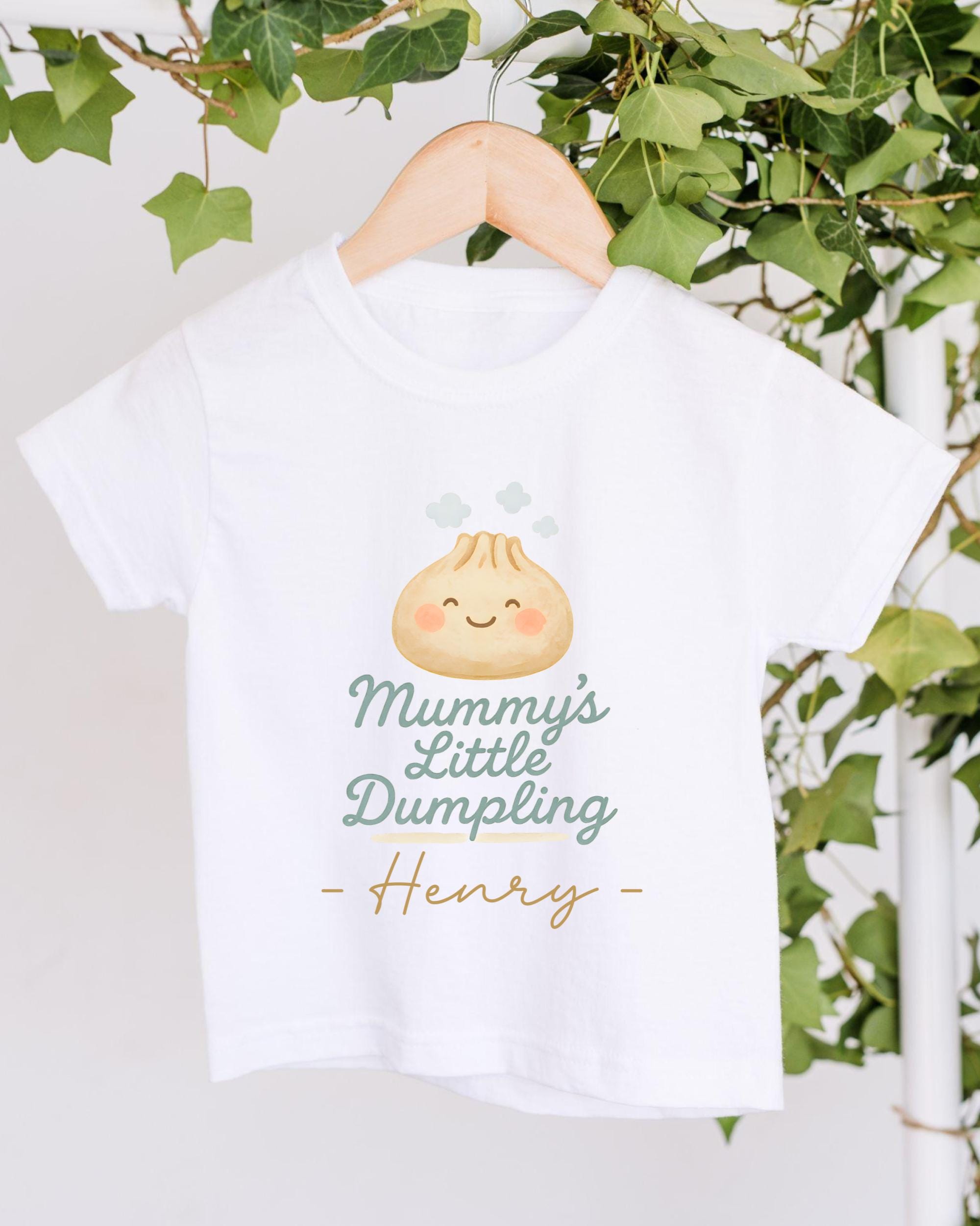 A white t-shirt with a cartoon image of a dumpling and the text "Mummy's Little Dumpling Henry" printed on it, hanging on a wooden hanger.
