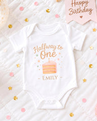 A white onesie with a birthday cake design and the text "Halfway to One - Emily" printed on it, surrounded by confetti and a "Happy Birthday" sign.
