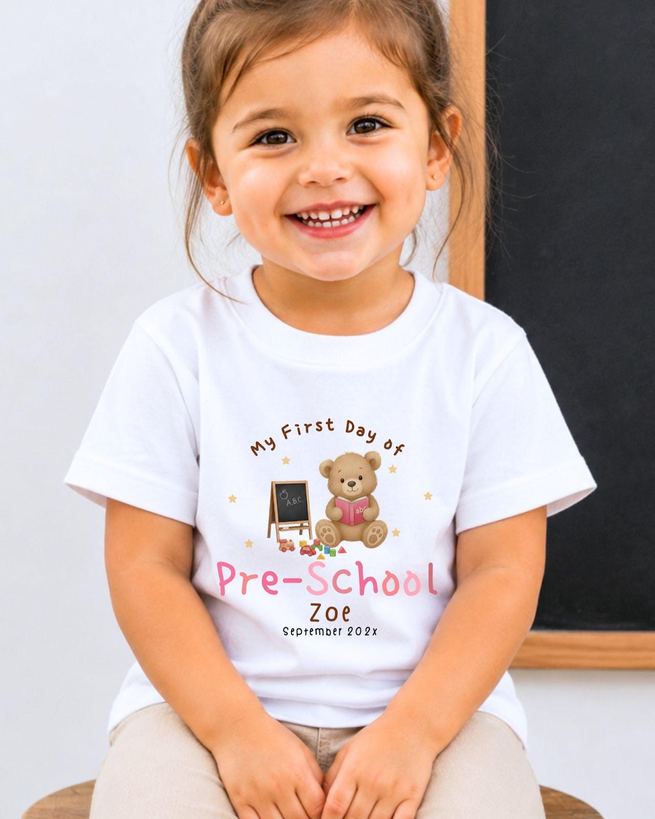 A young girl wearing a white t-shirt with a teddy bear and a chalkboard, smiling and sitting in front of a chalkboard.