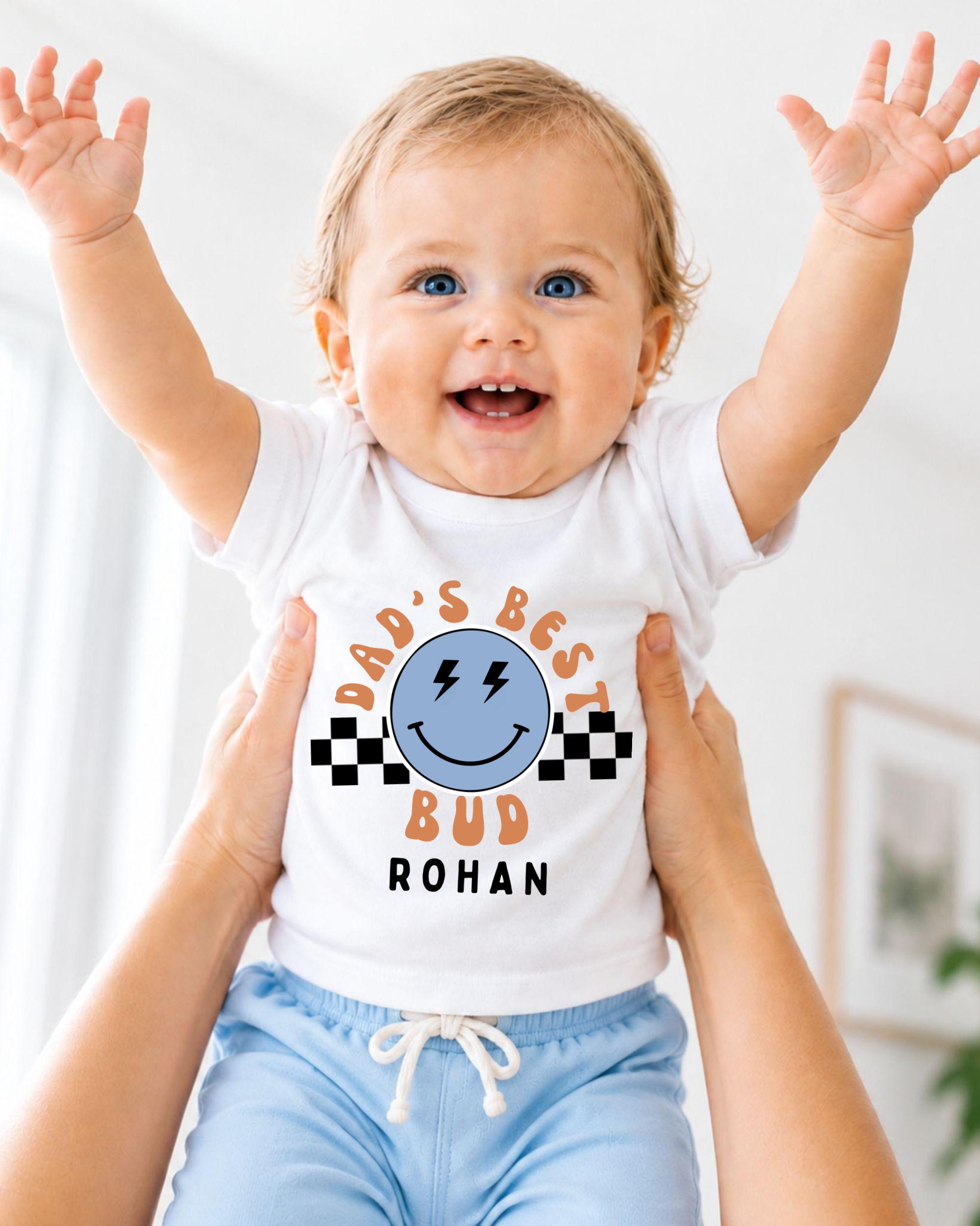 A smiling baby wearing a white t-shirt with a graphic design and text, being held up by someone's arms.